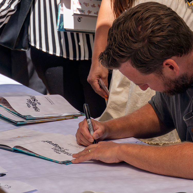 Man signing a book at an event with people in the background